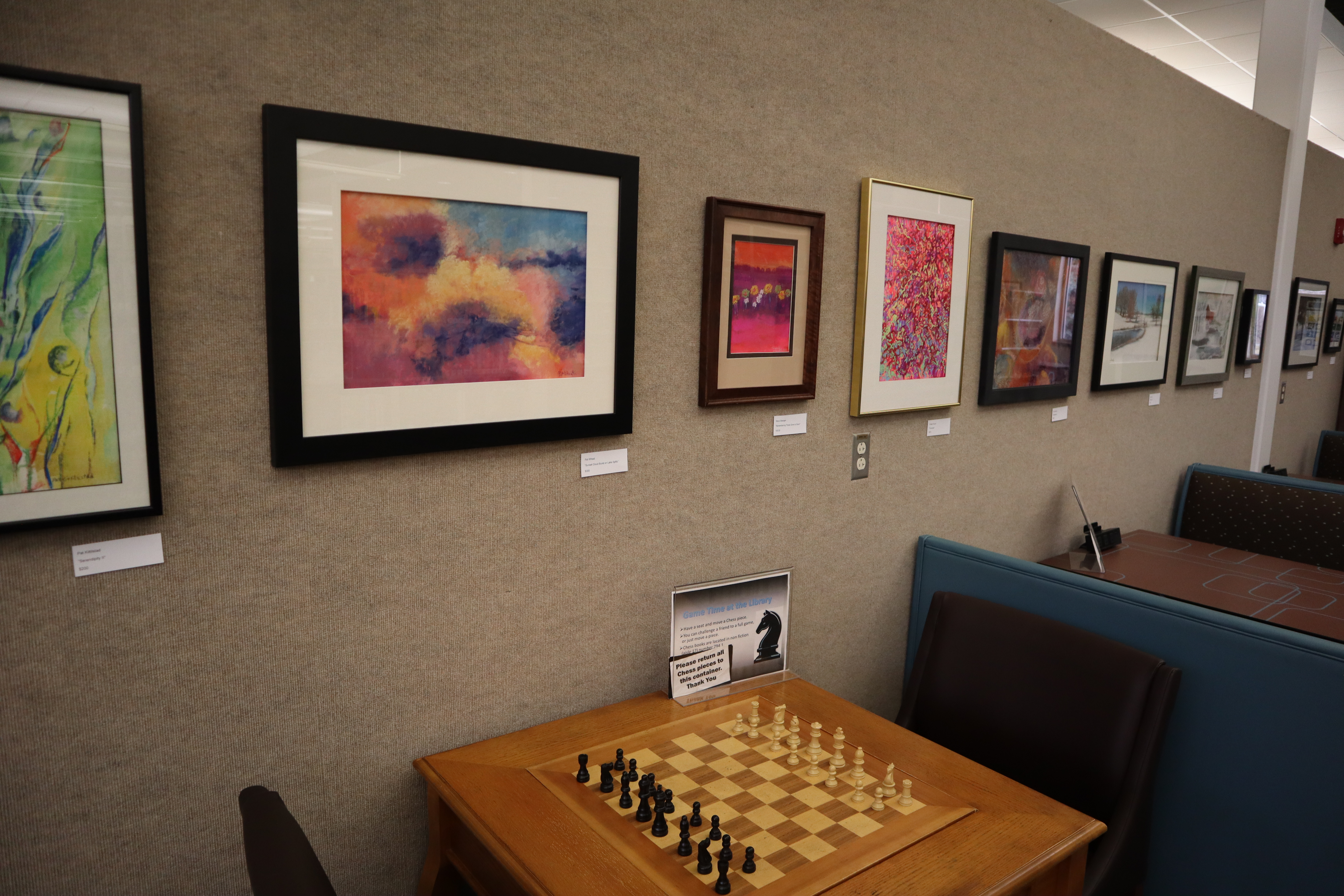 A long row of framed oil pastel paintings hanging on a wall above a seating area in the Library.