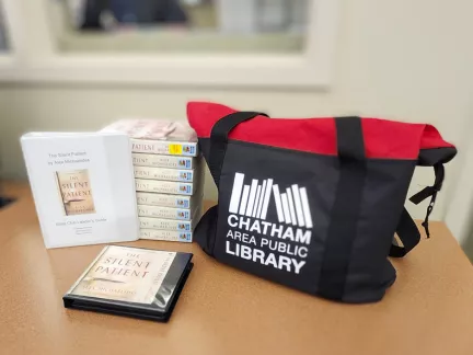 A bag containing several books is placed next to an open book on a table, inviting a moment of literary exploration.