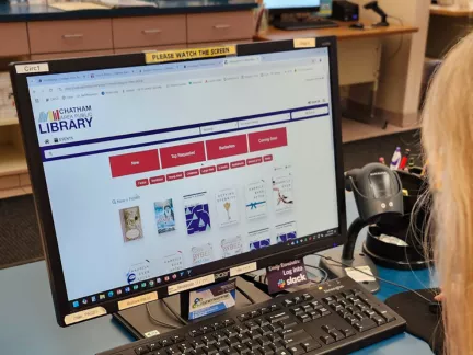 A woman sits at a computer in a library, focused on her work amidst books and quiet surroundings.